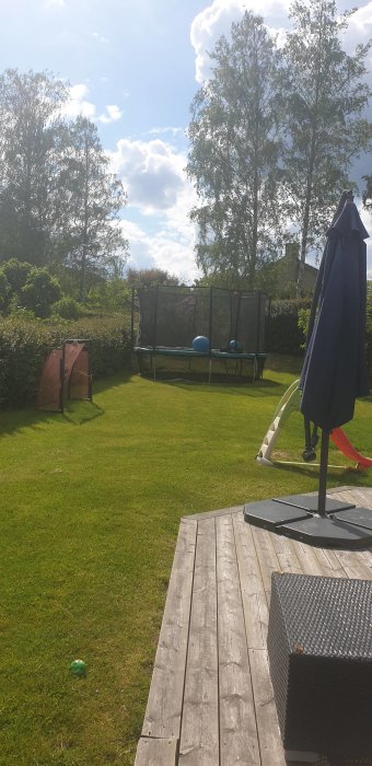 Solig trädgård med studsmatta, skyddsnät, gräsmatta, rutschkana och hopfällbart parasoll.