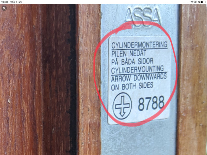 Instruktionsetikett för cylindermontage från ASSA på dörrkarm, riktning nedåt markerad.