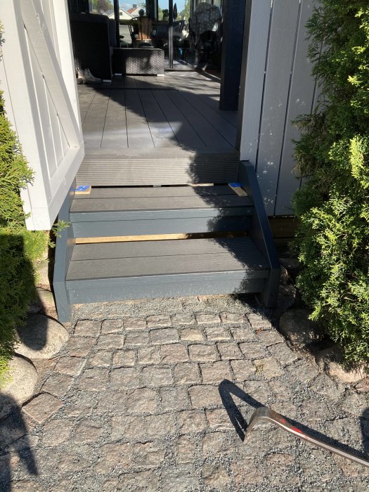 Trappa klädd med komposittrall som leder upp till en altan, omgiven av gröna buskar och stenläggning.