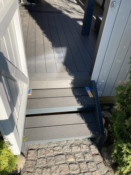 Trappa delvis klädd med komposittrall som leder upp till en altan, intill en vit husvägg och över stenläggning.