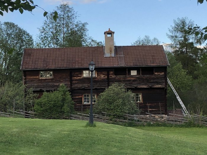 Mörkbrunt trähus med tegeltak och en inbyggd balkong på andra våningen till höger, omgivet av grönt gräs och träd.