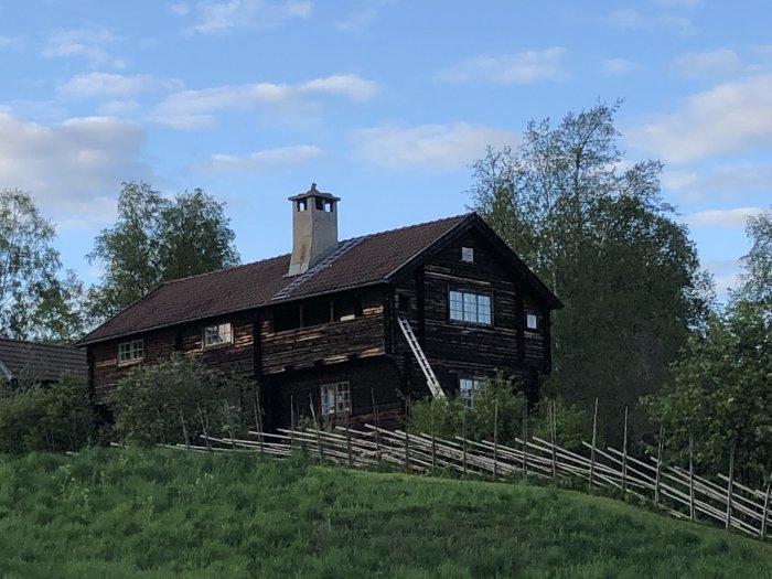 En mörkbrun trähusfasad med inbyggd balkong på övre våningen, omgiven av grönska och ett staket.