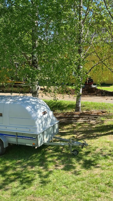 Släpvagn parkerad på gräsmatta med björkträd och uppgrävd mark i bakgrunden.