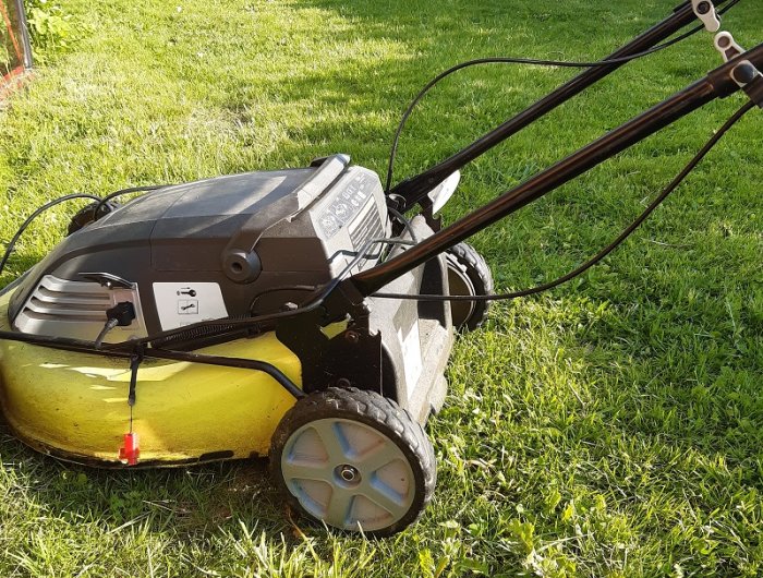 En Ryobi batteridriven gräsklippare på gräsmatta med fokus på bakhjulet och öppet handtag.