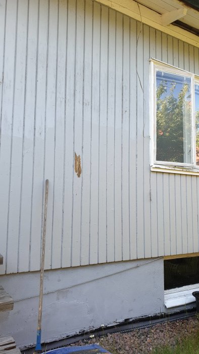Åldrad housefasad med flagande vit färg och synlig träyta vid den nedre delen, nära ett fönster.