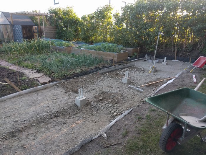 Grundarbete för byggprojekt med grus, betongfundament, skottkärra och trädgårdsredskap på en bakgård.