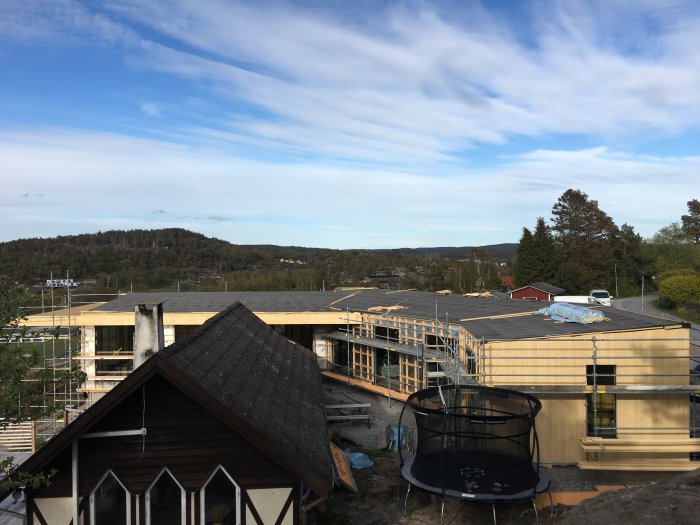 Pågående bygge av hus med uppsatt takstomme, fönsteröppningar och delvis påsatt träpanel, omgivet av byggnadsställningar.