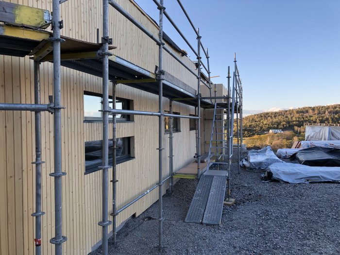 Byggnadsställning framför hus under konstruktion med träpaneler och fönster, detaljarbete syns.