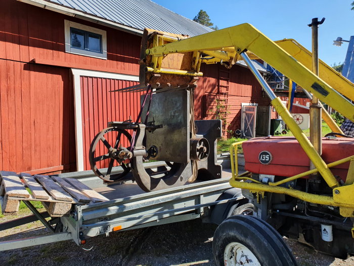 Bandsåg lastad på ett släp med hjälp av en gul, mobil kran, framför en röd lada och en röd traktor.