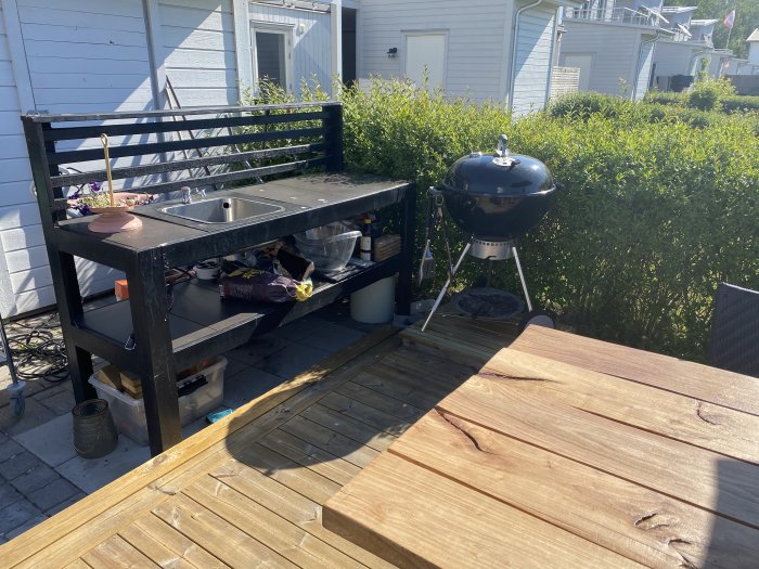 Hembyggt utekök med inbyggd diskho och ett handgjort trädgårdsbord bredvid en kolgrill på en träaltan.