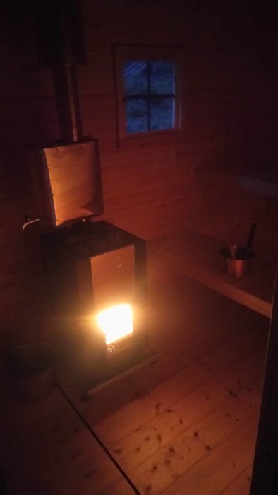 En bastu med upplyst kamin och mörkt trädgolv, en hink och en fönstervy i skymningen.