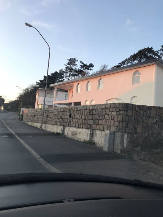 Naturstensmur framför ett rosa putsat hus sett från en bil på gatan vid skymningen.