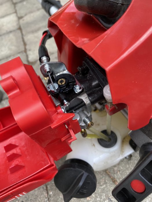 Närbild på en grästrimmers karburator och tändstift, synligt fuktigt av bensin, mot röd plastkåpa.