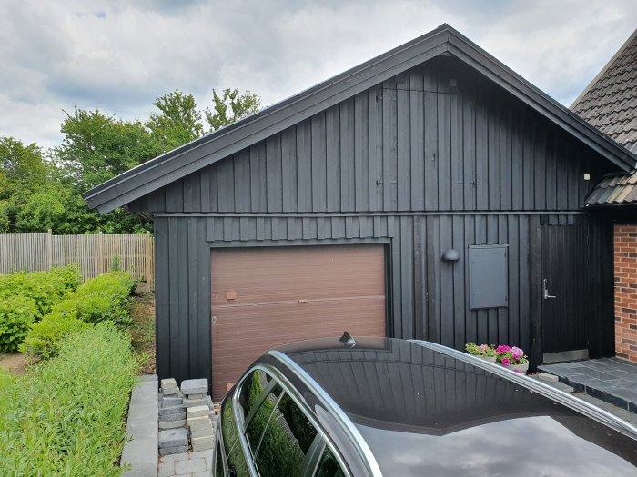 Svartmålat garage med sadeltak och garagedörr, intill bil och stenlagd väg.