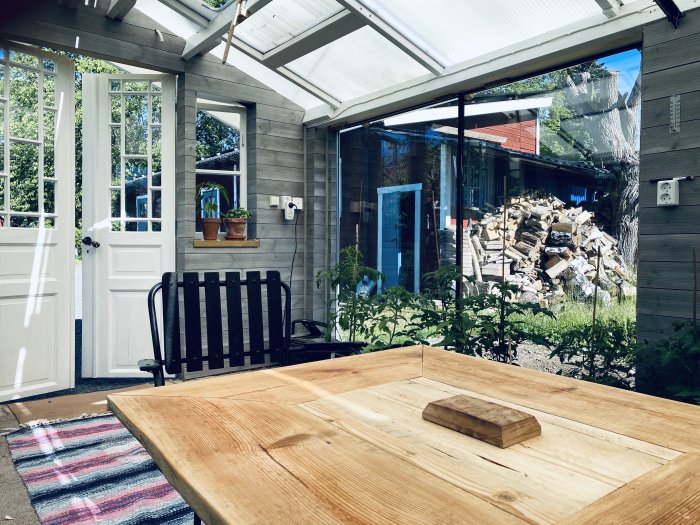 Hembyggt växthus med träbord, sittbänk och växtlighet, speglar ett kreativt byggprojekt med återanvänt material.