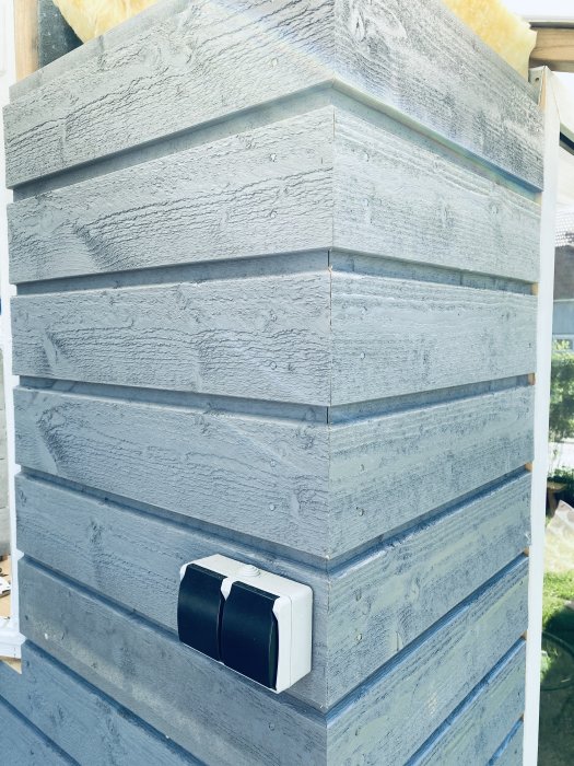 Vägg av blåmålat trä med horisontella paneler och en svartvit eluttagsbox, del av ett hemmabyggt växthus.