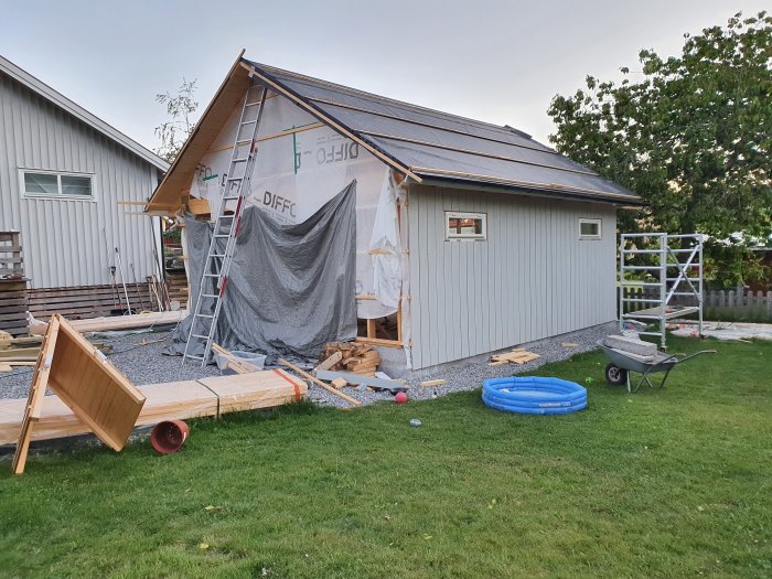 Ett garagebygge med material och byggverktyg utspridda framför, och uppspikad panel på ena långsidan.