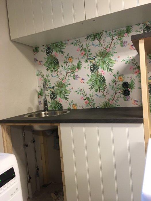 Oavslutad köksrenovering med blommig tapet, bänkskiva och synliga rör under diskbänk.