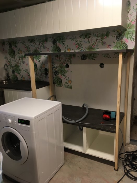 Rum under renovering med tvättmaskin, bänkskiva och överskåp mot blommig tapet.