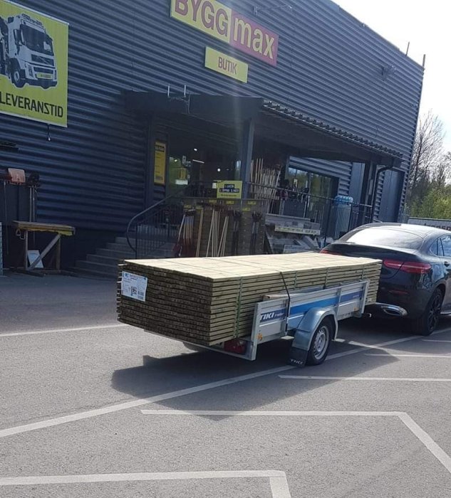 Överlastad släpvagn med träplank framför Byggmax, säkrad med endast ett spännband.