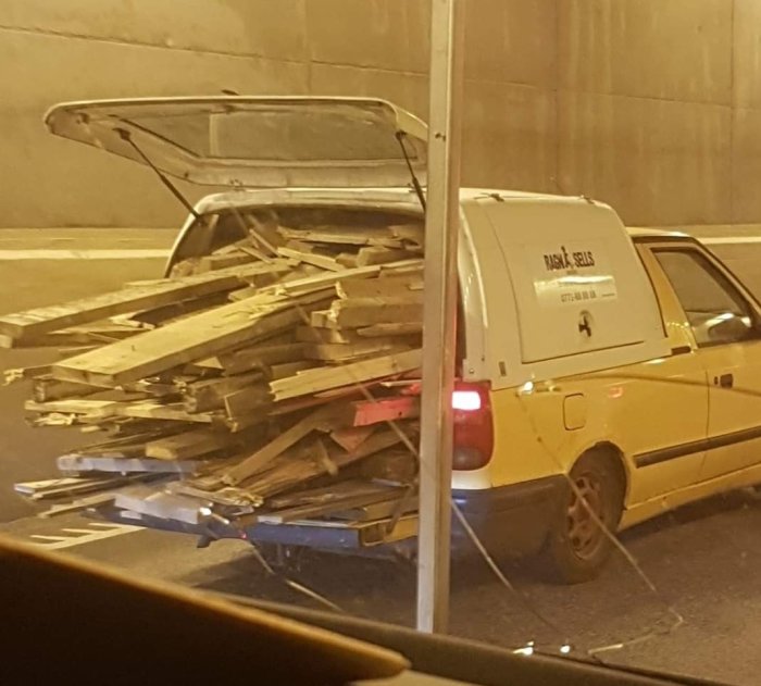 Överlastad gul skåpbil med bakluckan öppen och säkrad med ett spännband, full med byggbräder.