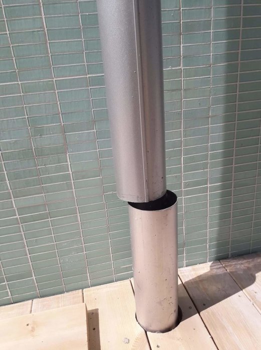 Grått avloppsrör som går genom en träplattform med grönt kakel i bakgrunden.