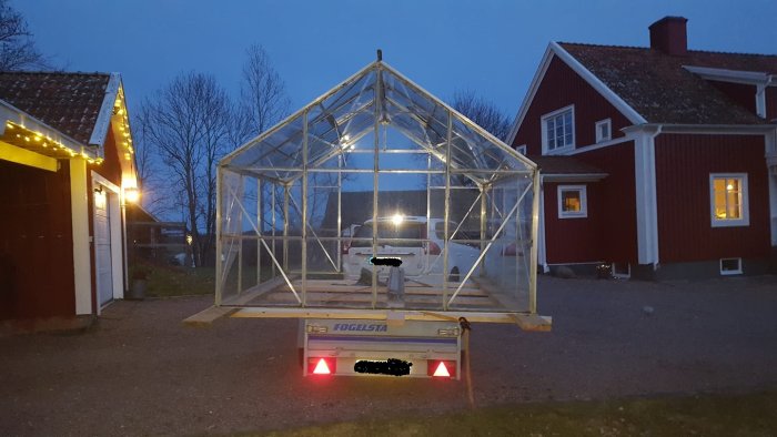 Trästomme till växthus monterad på släpvagn parkerad framför rödfärgat hus i skymningen.