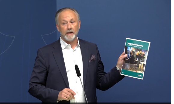 Man i kostym visar upp anslag med titeln "Safe to visit" under presskonferens.