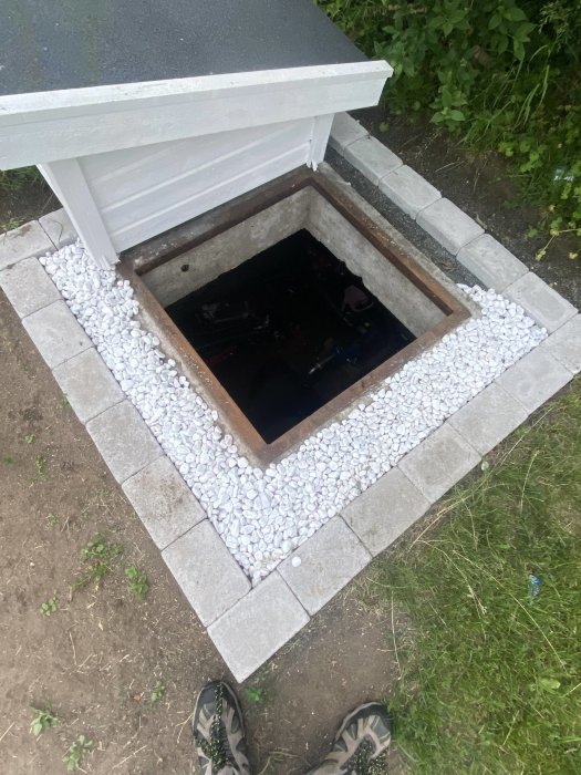 Öppen brunn med lock omgiven av vita stenar och grå trottoarplattor, synligt gräs och jord, stående person.