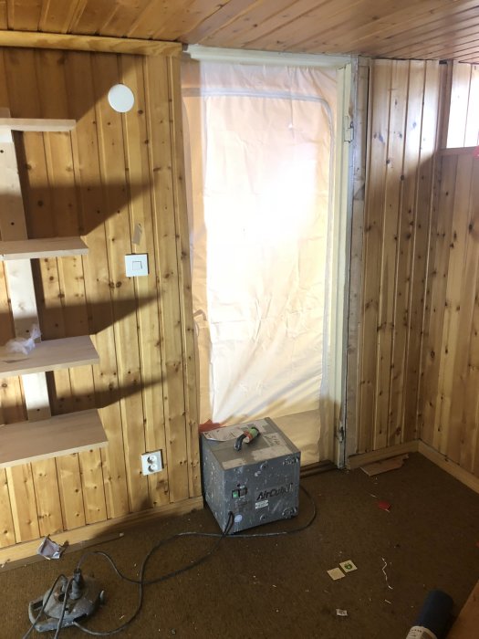 Renoveringsrum med träväggar och betonggolv i processen att omvandlas till förråd, med byggmaterial och verktyg synliga.