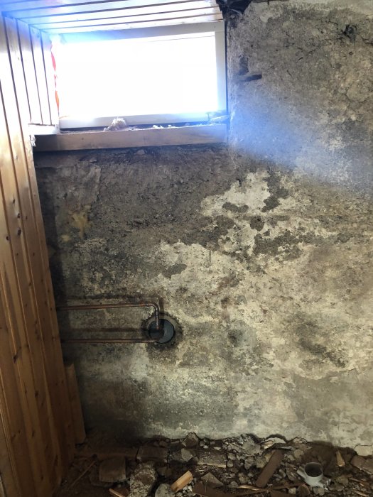 Rum under renovering med rå betongvägg, rörinstallation och öppet fönster som släpper in ljus.