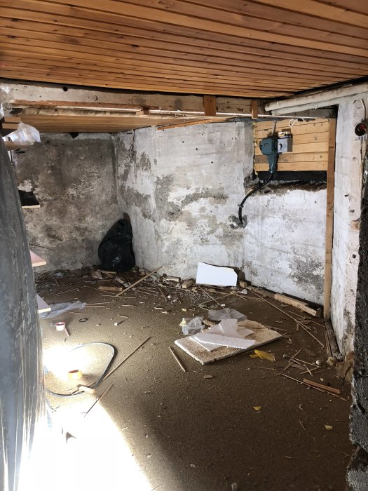 Oinrett förrådsrum under renovering med slitet betonggolv, skräp och träpanel i taket.