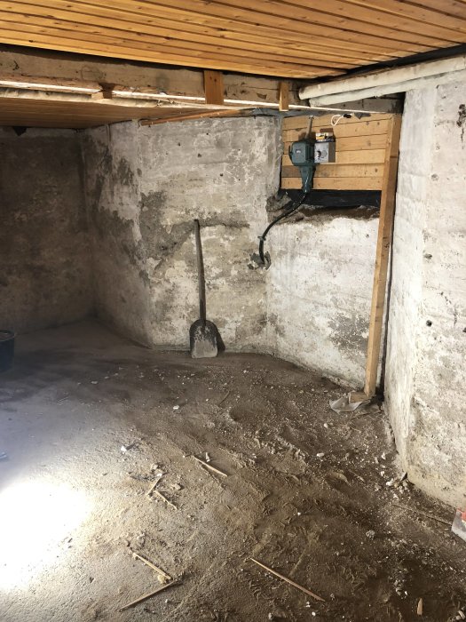 Ofärdigt förrådsrum med ojämnt betonggolv, spade och rörledningar under träbjälkar.