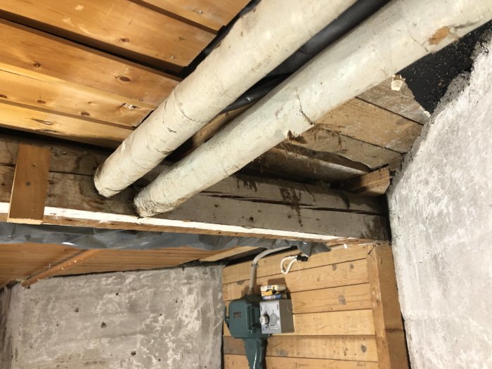 Översikt av ett ofärdigt rum med träbjälklag, betonggolv, och synliga rör under renovering för användning som förråd.