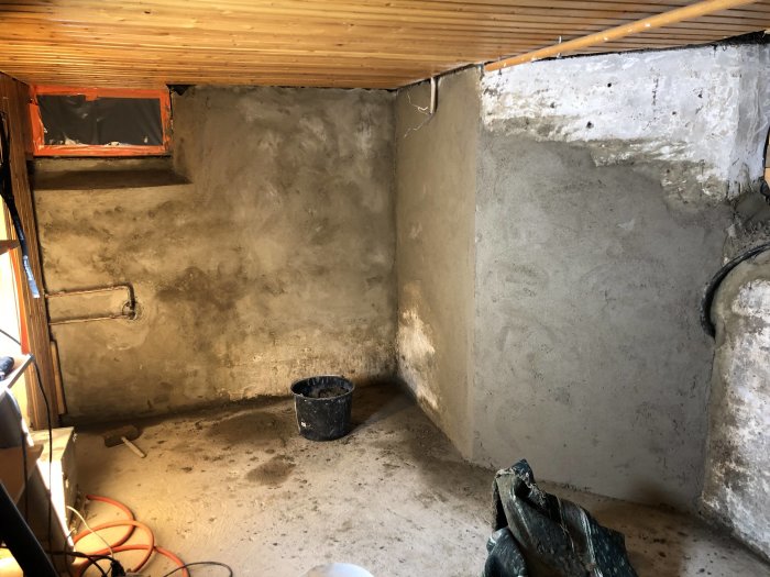 Ommålad källarvägg och golvrör, spackelhink och verktyg syns, med träträbjälklag ovanför.
