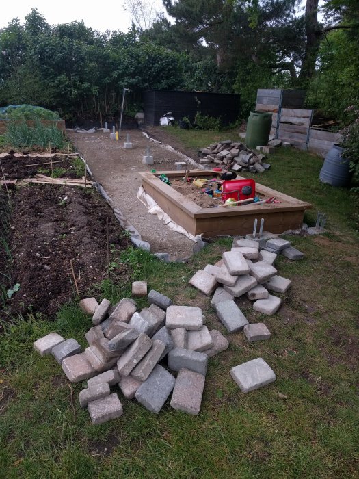 Hög med marksten i en trädgård för stenläggning, släpvagn och påbörjat projekt i bakgrunden.