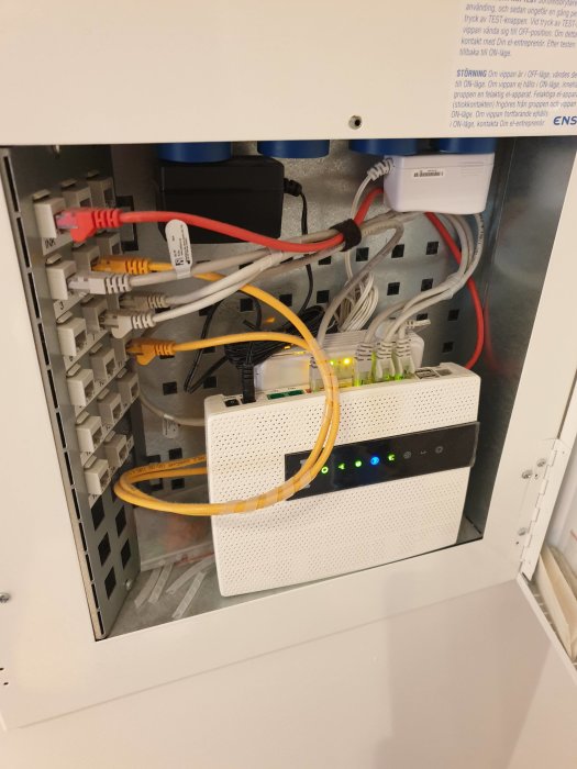 Öppet elskåp med nätverksutrustning och kablar, inklusive en switch med lysande indikatorlampor.