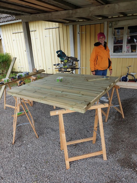 Person i orange jacka står bredvid nybyggt tryckimpregnerat golv på träarbetsbord utomhus.