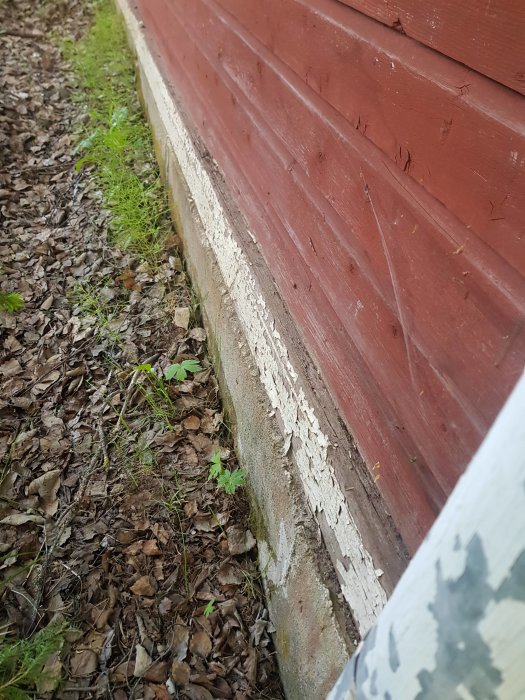 Nedre delen av ett rött trähus med skadad torpargrund där färg flagar och marken lutande, visar potentiella vattenskador.