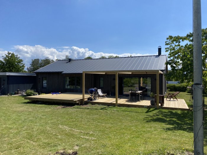 Trädgård med nästan färdigt svart trähus och altan under solig himmel, byggmaterial syns.