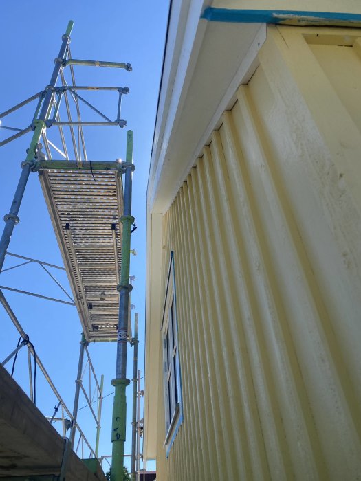 Byggnadsställning bredvid en gul husvägg med ny lockläkt under en klarblå himmel.