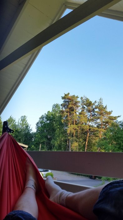 Person vilar i hängmatta på veranda med utsikt över träd och en bil i bakgrunden.