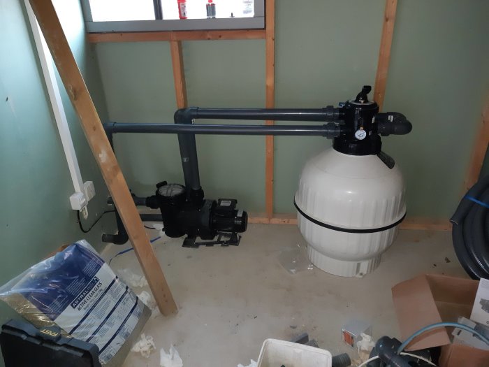 Installationsrum med nyligen limmade PVC-rör, en poolfiltertank och en pump, oordning på golvet.