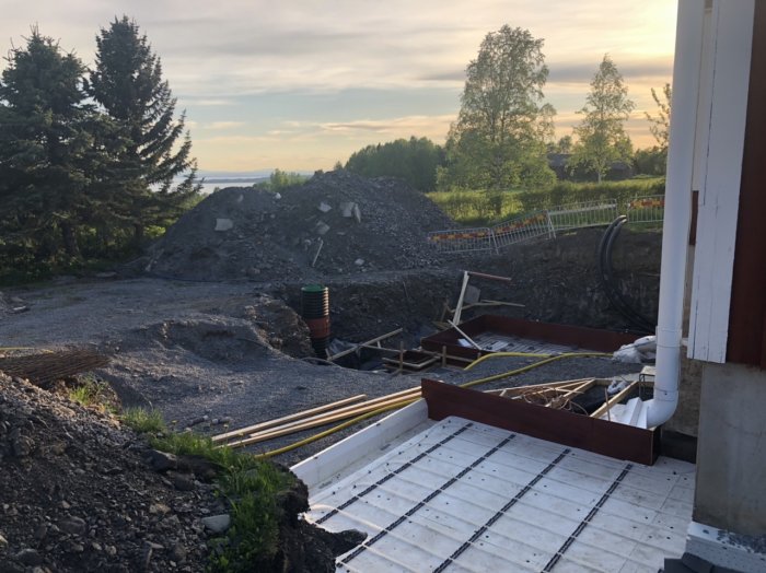 Stor grävd grop vid husgrund med byggmaterial, säkerhetsstaket och natur i bakgrunden.