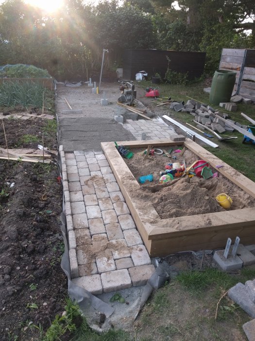 Pågående trädgårdsprojekt med delvis lagda grå trottoarstenar vid sidan av en sandlåda och byggmaterial spridda omkring.