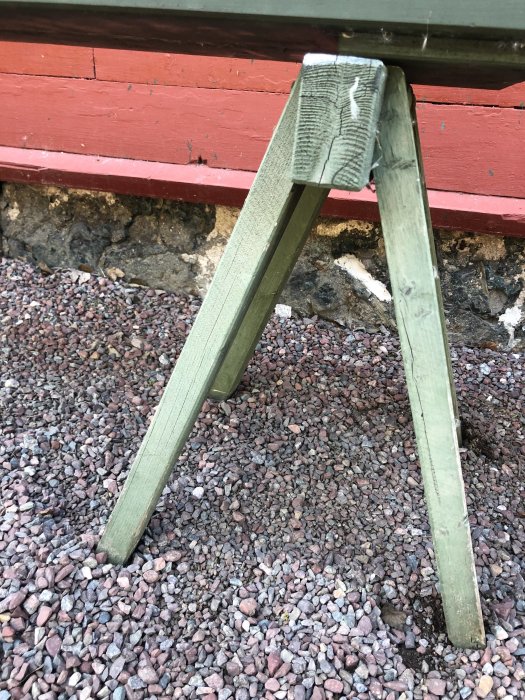 Nätt träbock med sammansatta ben under en överliggare, placerad på grus framför en rödmålad vägg.