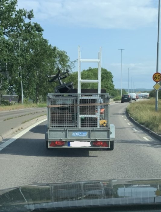 Släpvagn med byggmaterial och stege på en väg, sett från ett annat fordon.