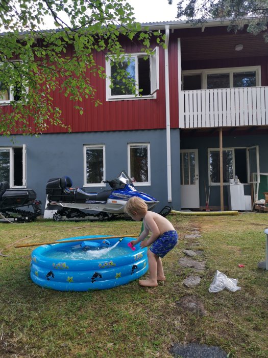 Barn fyller på vatten i en blå uppblåsbar pool framför en röd stuga med balkong.