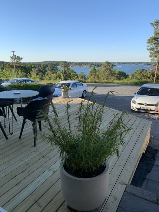 Nybyggd altan med bord, stolar och krukväxt framför vacker sjöutsikt, med bilar parkerade till höger.