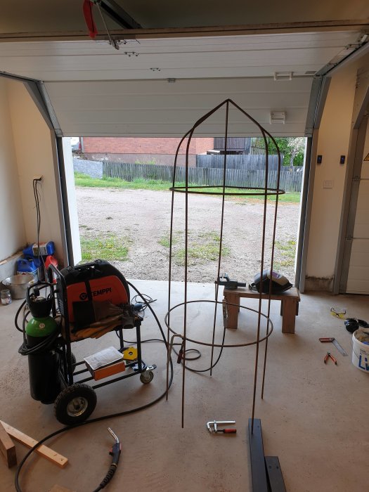 Hemmagjord rund båge av järn med fem lodräta stänger i ett garage, svetsutrustning i förgrunden.